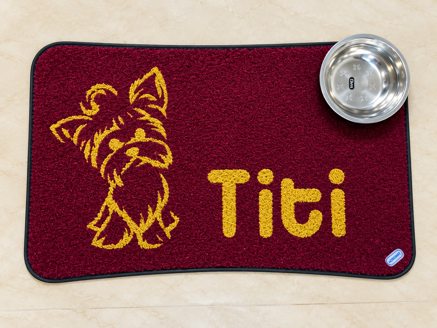 Tapete personalizado para mascotas de color vinotinto, con ilustración amarilla de un perro y el nombre “Titi”, acompañado de un comedero de acero inoxidable sobre un piso beige.